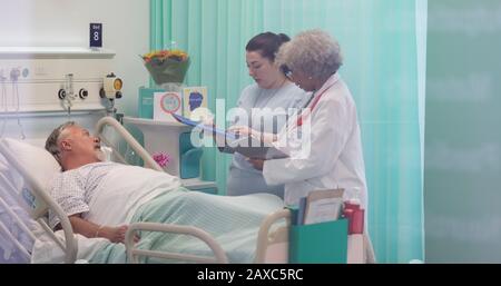 Ärzte mit medizinischen Diagrammen, die Runden drehen und mit dem leitenden Patienten im Krankenhauszimmer sprechen Stockfoto