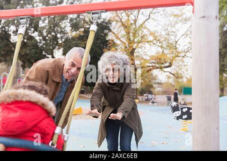 Verspielte Großeltern drängen Kleinkind Enkel auf dem Spielplatz auf Schaukel Stockfoto