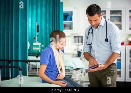 Kinderarzt zeigt digitale Tablette für Jungen mit Arm in Schlinge im Krankenhaus Stockfoto