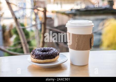 Kaffee in Papierbecher und Donuts mit Schokolade bestreut auf Plattenständer auf Holzbalkon. Snack im Urlaub mit Blick auf die Berge Stockfoto