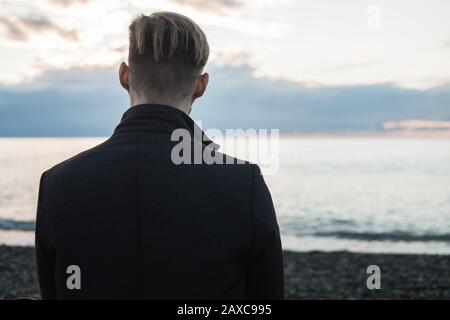 Junger männlicher Flusspferd in einem Mantel sitzt am Meer oder Ozean und genießt Sonnenuntergang. Herbst am Meer an einem felsigen Strand Stockfoto