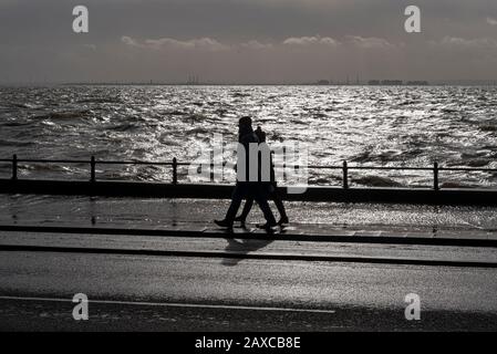 Männer- und Frauenpaar, die auf der Western Esplanade von Southend on Sea, Essex, Großbritannien spazieren und bei Sturmfluten des Sturms Ciara überflutete Straßenbeläge passieren Stockfoto