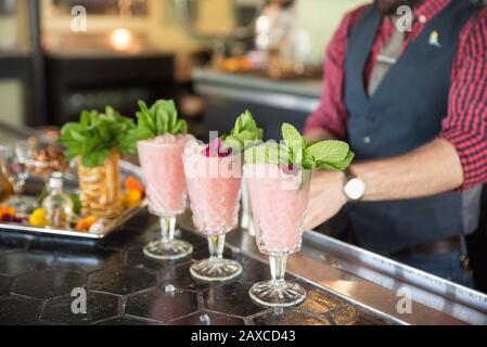 Drei rosafarbene Cocktails sitzen auf einem Bar-Top mit einem Tablett mit Zierspezialitäten in der Nähe und Barkeeper im Hintergrund. Stockfoto