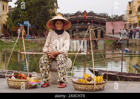 Alte vietnamesische Dame, die mit Obstkörben vor der japanischen Covered Bridge in Hoi An, Vietnam sitzt. Stockfoto