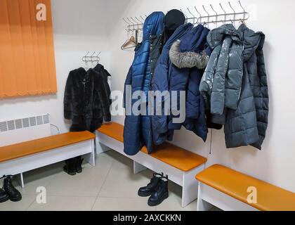 Jacken und Winteroberbekleidung hängen an Haken an der Wand. Im Umkleideraum der Frauen hängen Jacken, Fellmäntel und Mäntel. Orangefarbene Bänke, Schuhe und Klo Stockfoto