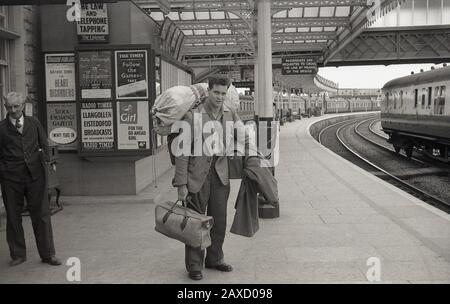 1950er Jahre, historisch, mit Taschen beladen, ein junger Mann, der auf einem Bahnsteig steht, möglicherweise nach Hause kommt, nachdem er seinen nationalen Dienst geleistet hat, daher die Kit-Tasche auf seinem Rücken, Weston-super-Stute, Somerset, England, Großbritannien. Auf den Postern des Senders finden Sie eine für die Radio Times, in der die Musiksendungen Llangollen & Eisteddfod, die Börsenzeitung Girl und eine für das Gesetz und das Telefonklopfen erwähnt werden. Stockfoto