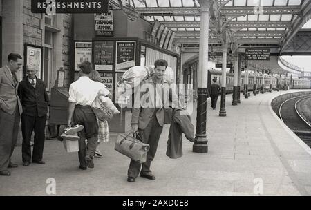 1950er Jahre, historisch, mit Taschen beladen, ein junger Mann, der auf einem Bahnsteig steht, möglicherweise nach Hause kommt, nachdem er seinen nationalen Dienst geleistet hat, daher die Kit-Tasche auf seinem Rücken, Weston-super-Stute, Somerset, England, Großbritannien. Ein Bahnhofsporter steht unter einem Bahnhofsschild für Erfrischungen. Auf den Plakaten ist eines für die Radio Times zu sehen, auf dem die Musiksendungen Llangollen & Eisteddfod, die Börsenzeitung, das Magazin Girl und eines mit dem Titel The Law and Telephone Tapping erwähnt werden. Stockfoto