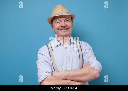 Porträt eines freundlichen, selbstbewussten Seniors im Sommerhut, der isoliert über blauem Hintergrund steht. Positivre Gesichtsemotion des Menschen. Stockfoto