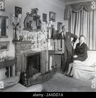 1950er Jahre, historisch, eine gut gekleidete Dame und Gentleman in ihrer eleganten Lounge, mit dekorativem Kamin und Kunstwerken an der Wand, Irland. Stockfoto