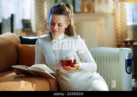 Trendige Frauen in weißem Pullover und Rock in der Nähe der Couch neben weißem Elektro-Ölkühler und Lesen mit Tasse heißen Tee im modernen Wohnzimmer Stockfoto