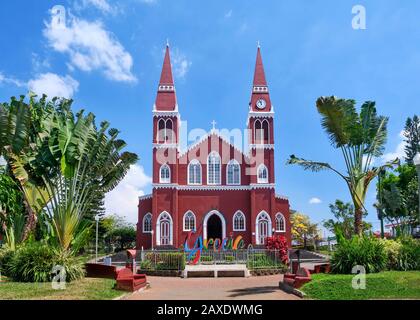 Die rot-weiß-weiße katholische Kirche in Grecia Costa Rica liegt zwischen grünen Palmen und unter einem wunderschönen blauen Himmel mit weißer flauschiger Wolke Stockfoto