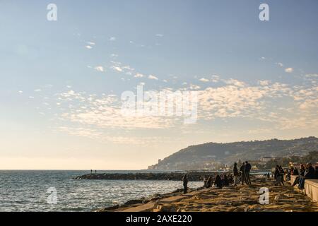 Menschen und Touristen genießen die Sonne auf den Felsen der Küste in einem warmen Wintertag mit Capo Nero cape im Hintergrund, Sanremo, Ligurien, Italien Stockfoto