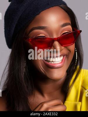 Nahaufnahme des Porträts einer jungen afrikanischen Dame, die positiv lacht und schöne Zähne zeigt und in einem Studio auf grauem Hintergrund einen Hut mit Brille trägt Stockfoto