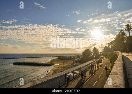 Hintergrundbeleuchteter, malerischer Blick auf die Küste mit Menschen und Touristen, die auf dem Fußgänger- und Radweg am Ufer des Meeres, Ligurien, Italien, spazieren gehen und radeln Stockfoto