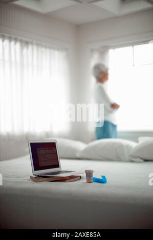 Öffnen Sie den Laptop, zwei Bücher, eine Tablettenflasche und einen Medikamentenspender, der auf einem Bett sitzt und eine Frau vor einem Fenster im Hintergrund steht. Stockfoto