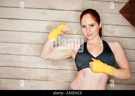 Junge Frau in einem schwarzen Sport-BH und gelben Boxhandwicklungen zerseigt stolz eine muskulöse Wange, während sie für ein Porträt vor einer Holzwand posiert. Stockfoto