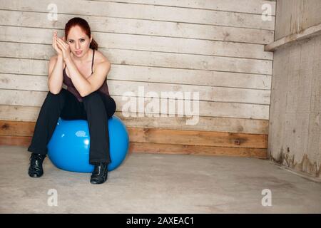 Junge Frau mit muskulösen Armen sitzt auf einem blauen Übungsball, der ihre Ellbogen auf die Knie lehnt und ihre Hände von ihrer Wange umklammert, wie sie für ein Porträt aufwirft. Stockfoto