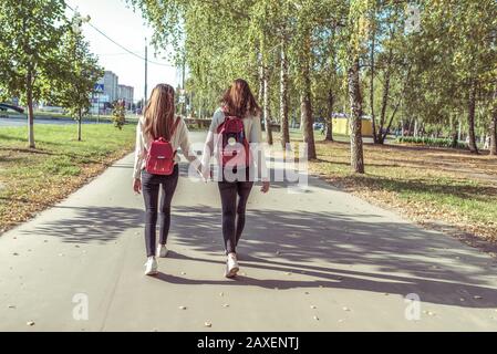 Zwei Mädchen Freundinnen Schulmädchen gehen im Sommerpark, Hintergrund Straßenbäume, Blick von hinten, Rucksäcke dahinter, Rückkehr nach der Schule, Straße nach Hause Stockfoto