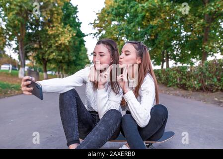 Zwei Mädchen Schüler 12-14 Jahre alt, im Sommer im Stadtpark, fotografieren selfie auf Smartphone, sitzen auf Skateboard, legere Kleidung, Pausenschule Stockfoto