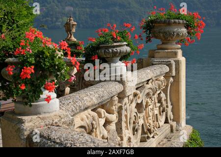 Villa del Balbianello Rotes Geranium in Urnen an einer verzierten Terrassenwand, Cherubs, Akanthuswirbel, Blick auf den Comer See, Lombardei Italien,
