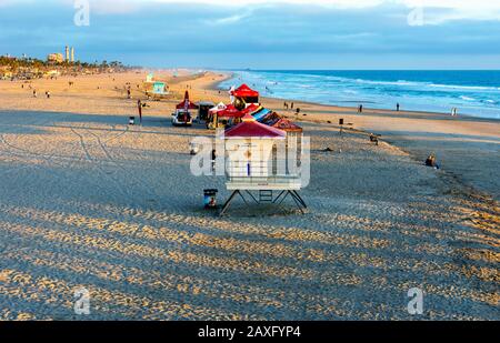 Blick auf den Strand, den Pazifischen Ozean, einen Rettungsschwimmturm und die Skyline der Stadt vom Huntington Beach Pier aus, während die untergehende Sonne Schatten erzeugt. Stockfoto