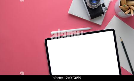 Overhead-Aufnahme eines eleganten Designer-Arbeitsbereichs mit verspottem Tablet, Kamera, Briefpapier und Kopierbereich auf pinkfarbenem Tischhintergrund Stockfoto