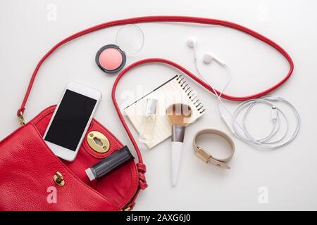 Rote Frauentasche mit Kosmetik, Parfüm, Armband, Smartphone, Ohrhörer und Notizblock auf weißem Hintergrund, Draufsicht Stockfoto