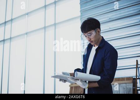 Asian man Carpenter arbeitet mit technischem Zeichnen oder Baupapier, das auf einer Werkstatt mit Zimmermannswerkzeugen und Holz zu Hause liegt Stockfoto