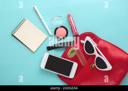 Modetasche aus rotem Leder mit Lippenstift, Pinsel, Notizblock, Stift, Smart Watch, Sonnenbrille, Smartphone auf blauem Hintergrund, flacher Lay Stockfoto
