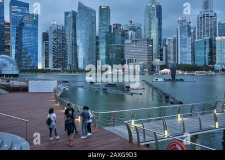 In der Abenddämmerung an einem grauen Tag in Marina Bay, Singapur, genießt sich eine Gruppe junger Touristen; b/g: Die hohen Anstiege des Bankenviertels Stockfoto