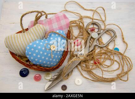 Ein Korb voller Herzen, genäht aus Stofffetzen, neben großen Scheren des Retro-Schneiders, grobem Faden und bunten Knöpfen. Stockfoto