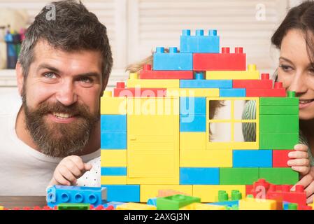 Das Kind versteckt sich hinter dem Haus aus Plastikblöcken. Familie auf unschärfem hellem Hintergrund. Mutter und Vater in der Nähe von Spielzeughaus aus bunten Ziegelsteinen. Familienspielkonzept. Stockfoto