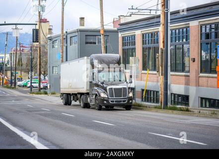 Dunkelbrauner Big Rig Day CAB-Sattelschlepper mit Dachspoiler, der Waren in einem kurzen trockenen Transporter transportiert, um die Manövrierbarkeit beim bergauf fahren zu verbessern Stockfoto