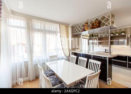 Weißer rechteckiger Tisch mit weißen Stühlen mit hohen Rückstützen im Küchenwohnzimmer des Laksheri-Cottages im klassischen Stil. Acht Stühle, große Fenster Stockfoto