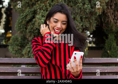 Attraktive junge Frau in Rot gekleidet, sitzt auf einer Bank im Park und schaut auf ihr Handy, lächelt und feiert gute Nachrichten. Teenager modern Stockfoto