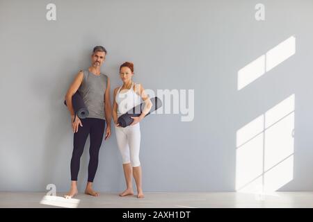 Yoga-Leute. Ein Mann und eine Frau mit Yogamatten stehen in einem hellen Klassenzimmer auf grauem Hintergrund. Stockfoto