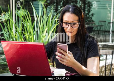Geschäftsfrau mit Brille, die eine Nachricht auf dem Handy textet, während sie ihren roten Laptop überprüft Stockfoto