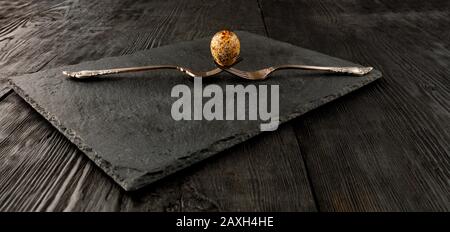 Wachtelei auf zwei alten gekreuzten Gabeln auf einer Küchenschneideplatte in schwarz aus Schiefer und einem dunklen Holzhintergrund. Taste "niedrig". Abgewinkelte Ansicht. Stockfoto