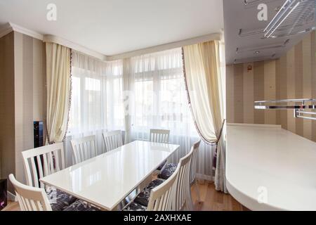 Weißer rechteckiger Tisch mit weißen Stühlen mit hohen Rückstützen im Küchenwohnzimmer des Laksheri-Cottages im klassischen Stil. Acht Stühle, große Fenster Stockfoto