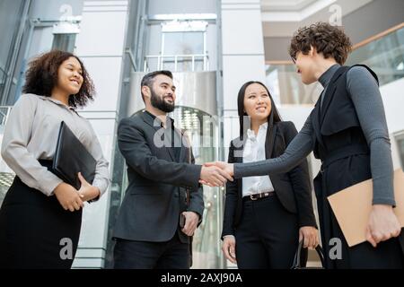 Lockiger Manager mit Ordnern, die die Hand des Mannes schütteln, während er mit dem Geschäftsteam im Büro zusammenkam Stockfoto