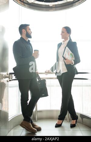 Positive junge Geschäftskollegen in formellen Outfits plaudern im gläsernen Aufzug Stockfoto