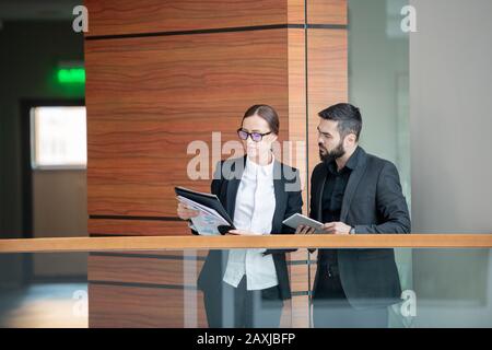 Nachdenkliche junge Business-Experten in Anzügen stehen am Geländer und prüfen gemeinsam Dokumente Stockfoto