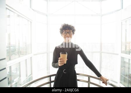 Portrait einer lächelnden, schönen jungen Dame mit lockigen Haaren, die die Kaffeetasse im glasigen Aufzug hält Stockfoto