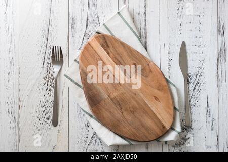 Ovales Schneidebrett mit Messer und Gabel mit Geschirrtuch auf weißem Holztischhintergrund Stockfoto