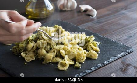 Nahaufnahme der Hand einer Frau, die mit einer glätterigen Gabel mit Brokkoli, einem typischen apulischen Rezept, in Hintergrundbeleuchtung auf dunklem Holztisch mit aufhebt Stockfoto