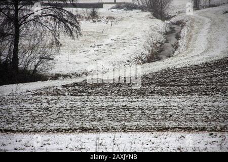 Felder mit einer dünnen Schneedecke neben einem Weg, Bach und Bäumen, winterlicher Stimmung, landwirtschaftliches Winterland im Süden des burgenländischen Stockfoto