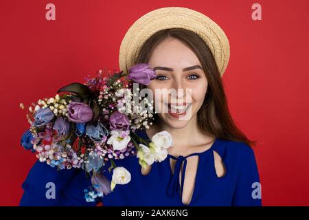 Fröhliche junge Frau in Hut, die Tulpenstrauß isoliert auf rotem Hintergrund hält. Stockfoto