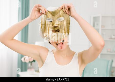Kosmetisches Verfahren, ziemlich lustige Mädchen in weißem Handtuch mit goldener Blattmaske auf hellem Hintergrund. Schönheits-, Spa-, Haut- und Körperpflege. Stockfoto