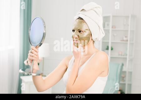 Gesichtshautpflege und Schönheitsbehandlungen. Frau mit einem Blatt, das die Goldmaske auf dem Gesicht feuchtet, und einem weißen Handtuch auf dem Kopf, mit Blick auf den Spiegel. Stockfoto