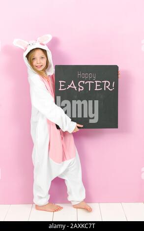 Hübsches blondes Mädchen mit gemütlichem Kaninchen-Kostüm und Tafel mit den Worten Happy easter posiert im Studio und ist glücklich Stockfoto